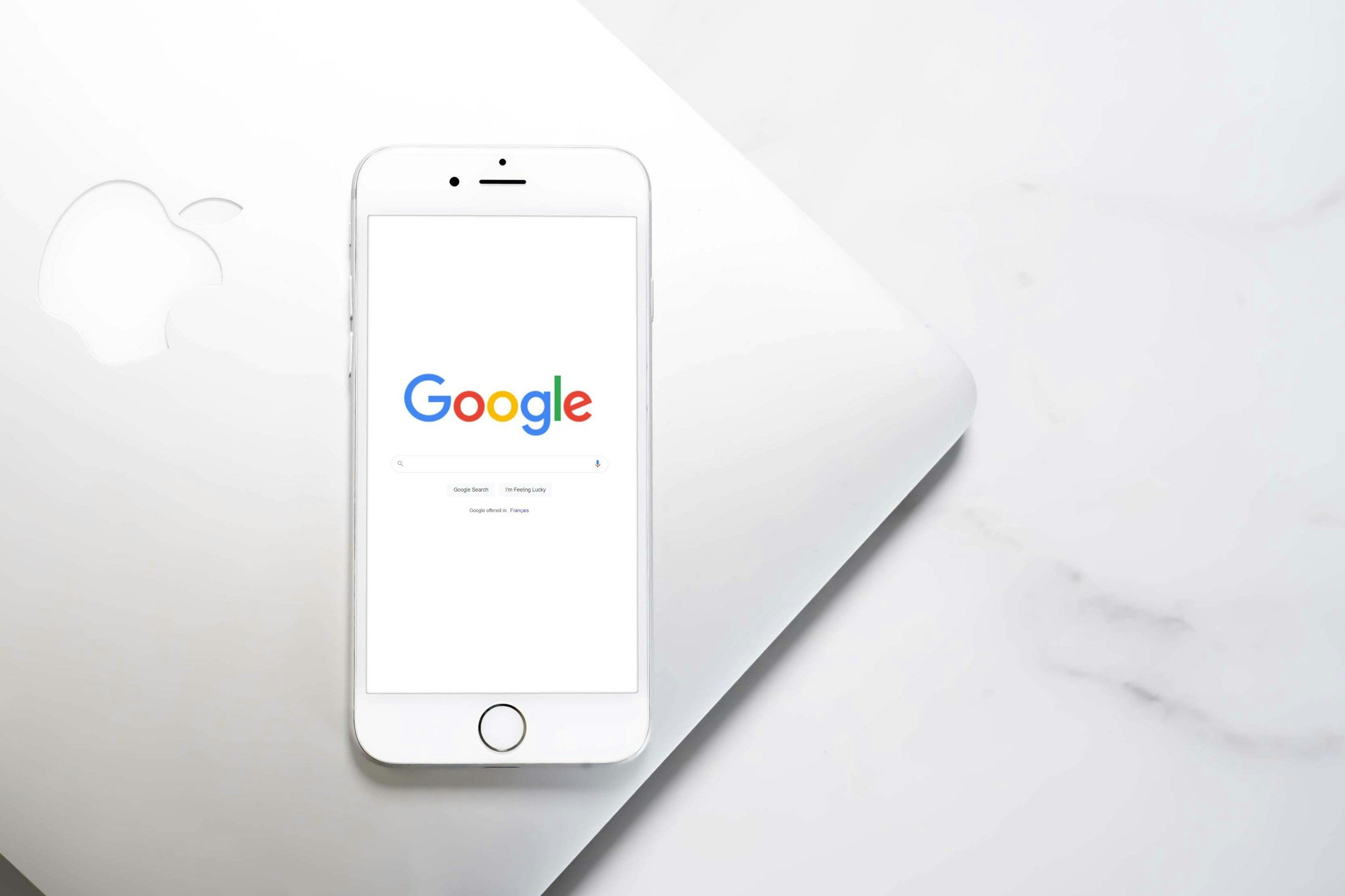 An iPhone displaying the Google search homepage on a white surface with a faint Apple logo in the background.