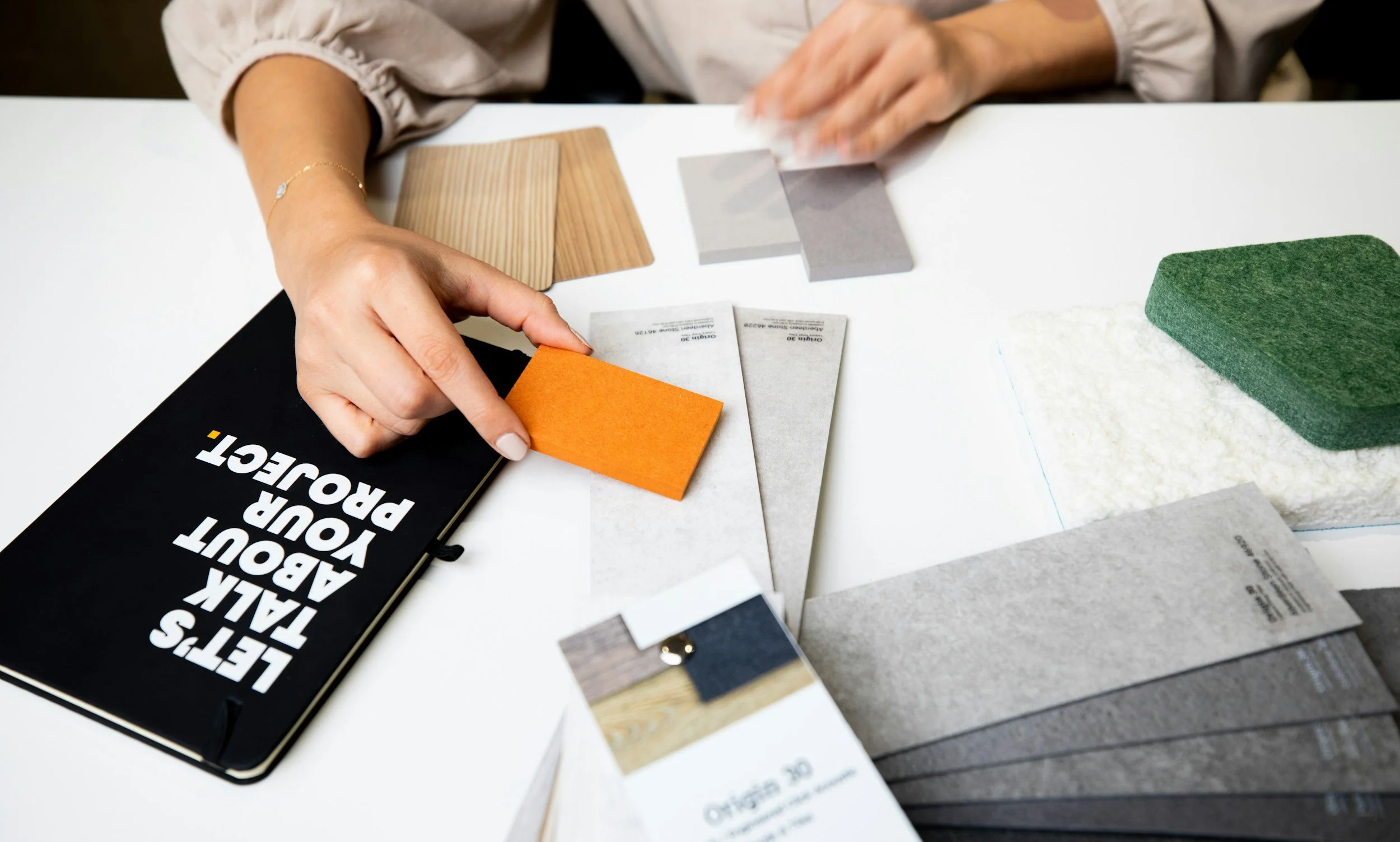 A person working at a table with various material samples including tiles, paint swatches, and a book titled 'Let's Talk About Your Project'.