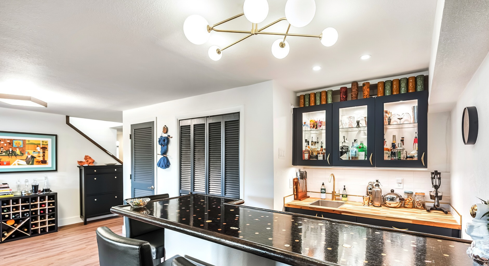 Modern kitchen with a black granite countertop island, black cabinetry with glass doors, a wine rack, and a bar area with bottles. Decor includes colorful jars on top of cabinets and a wall clock, with contemporary lighting fixtures.