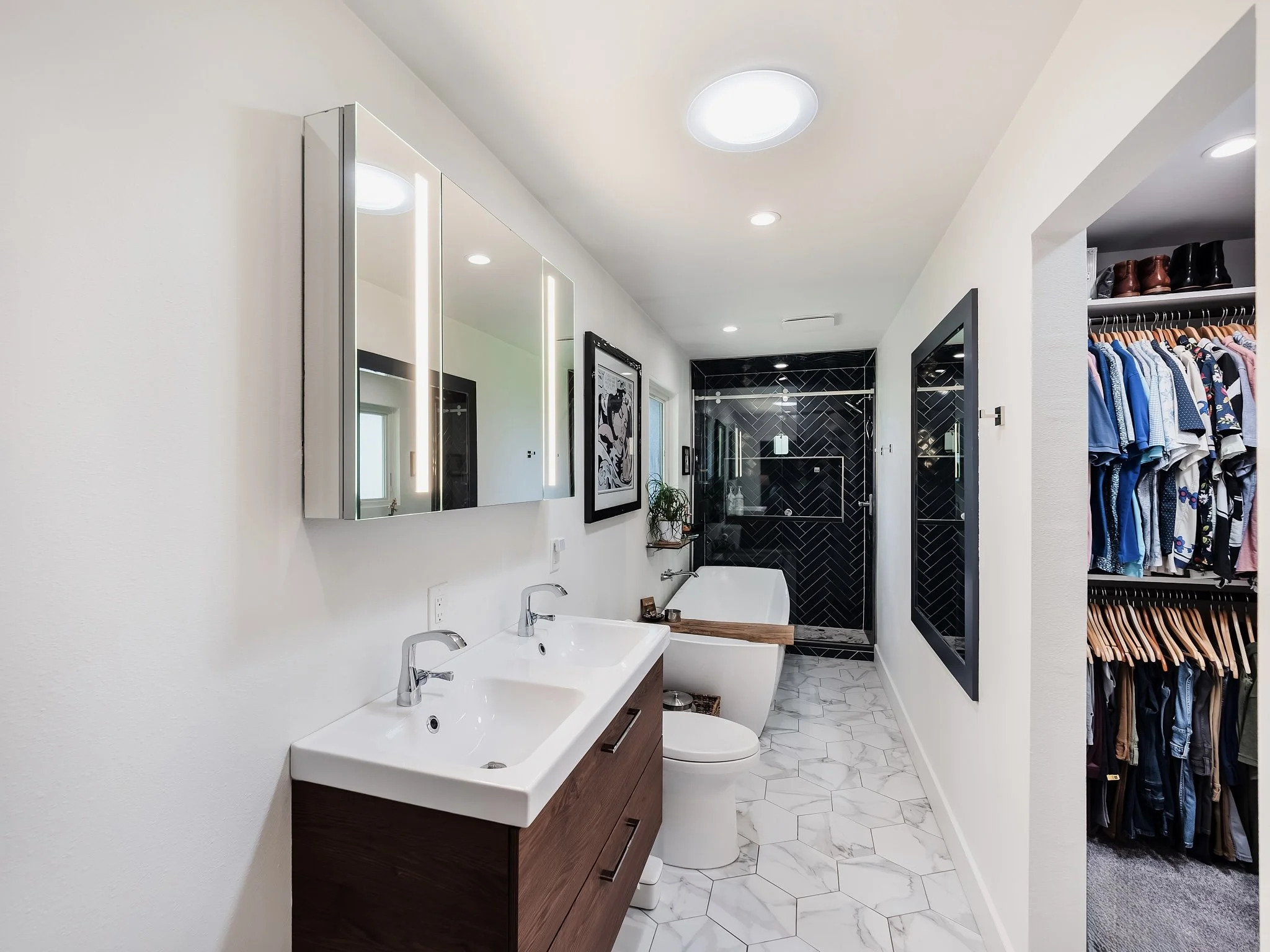 Modern bathroom with white walls, a double sink vanity with a wooden cabinet, a white bathtub, a black-tiled shower, and a walk-in closet with a row of hanging clothes and shoes.