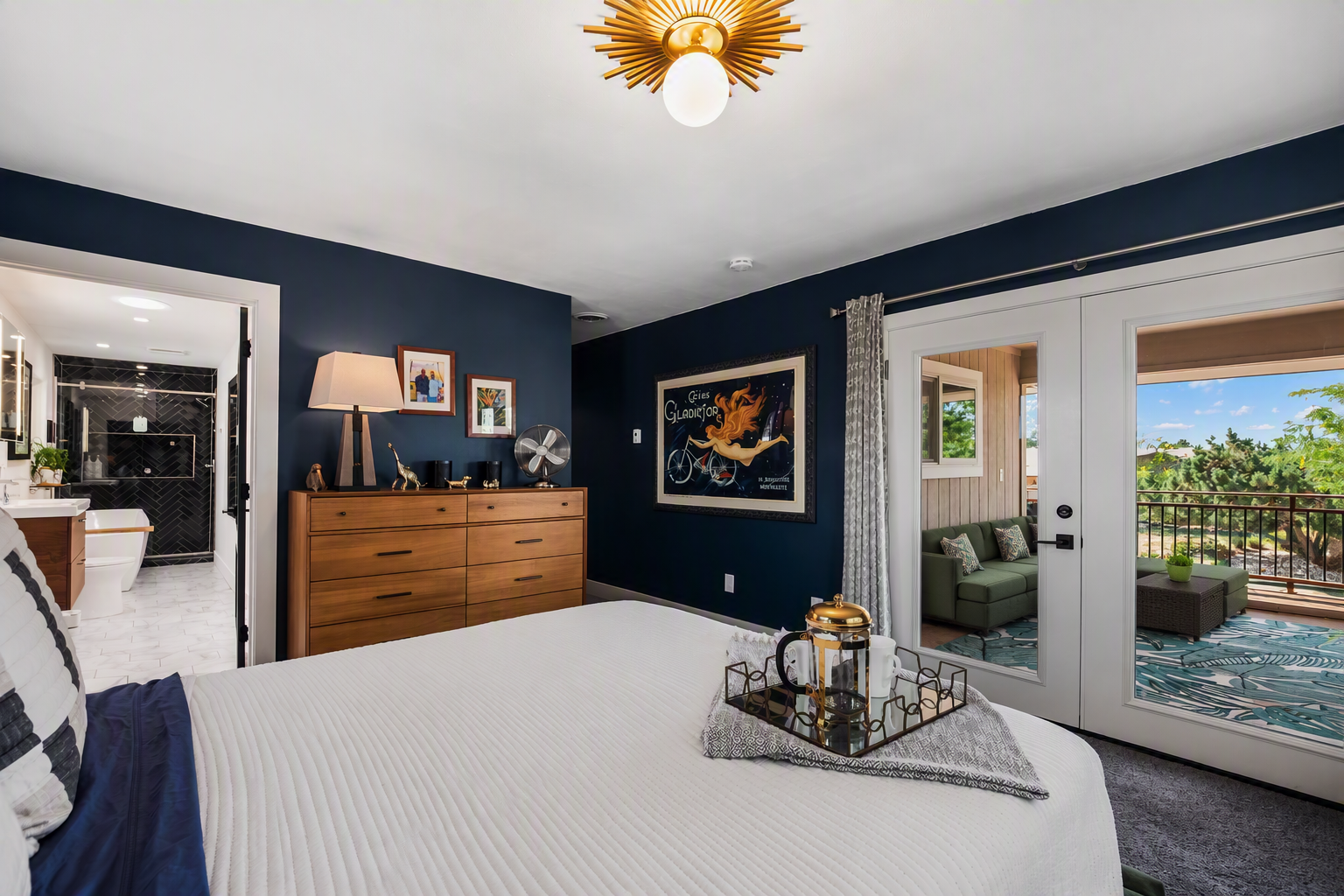 A bedroom with a white bed, a wooden dresser, and decor including framed pictures, a lamp, and a fan. There's a colorful poster on the dark blue wall. French doors open to a balcony with outdoor seating and a view of trees and a partly cloudy sky.