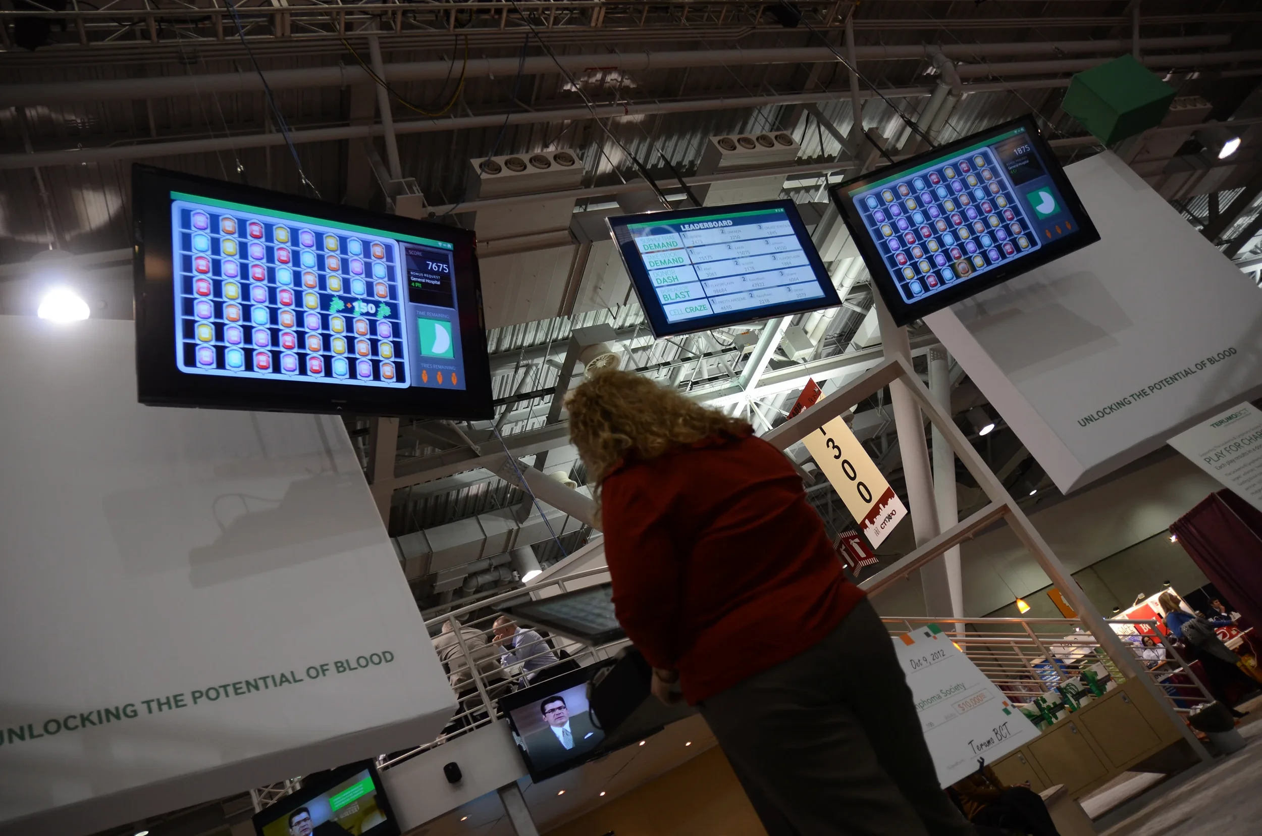 A woman playing a custom video game on a screen at conference, with two other screens show a leaderboard, and informational signs about blood donation in the background.