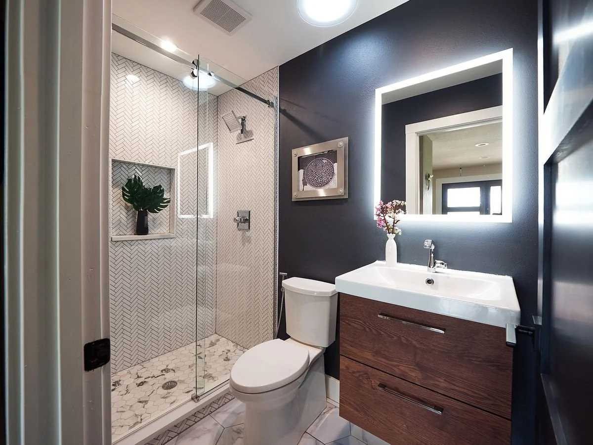 Modern bathroom with walk-in shower, white hexagonal floor tiles, dark wall, mirror with backlight, wooden vanity, and a small window.