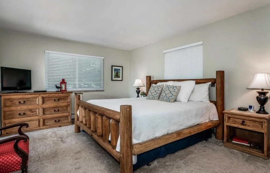 A bedroom with a wooden bed frame, white bedding, two decorative pillows, two nightstands with lamps, a dresser with a TV and red decorative item, a small framed picture, two windows with blinds, and a chair with red upholstery.