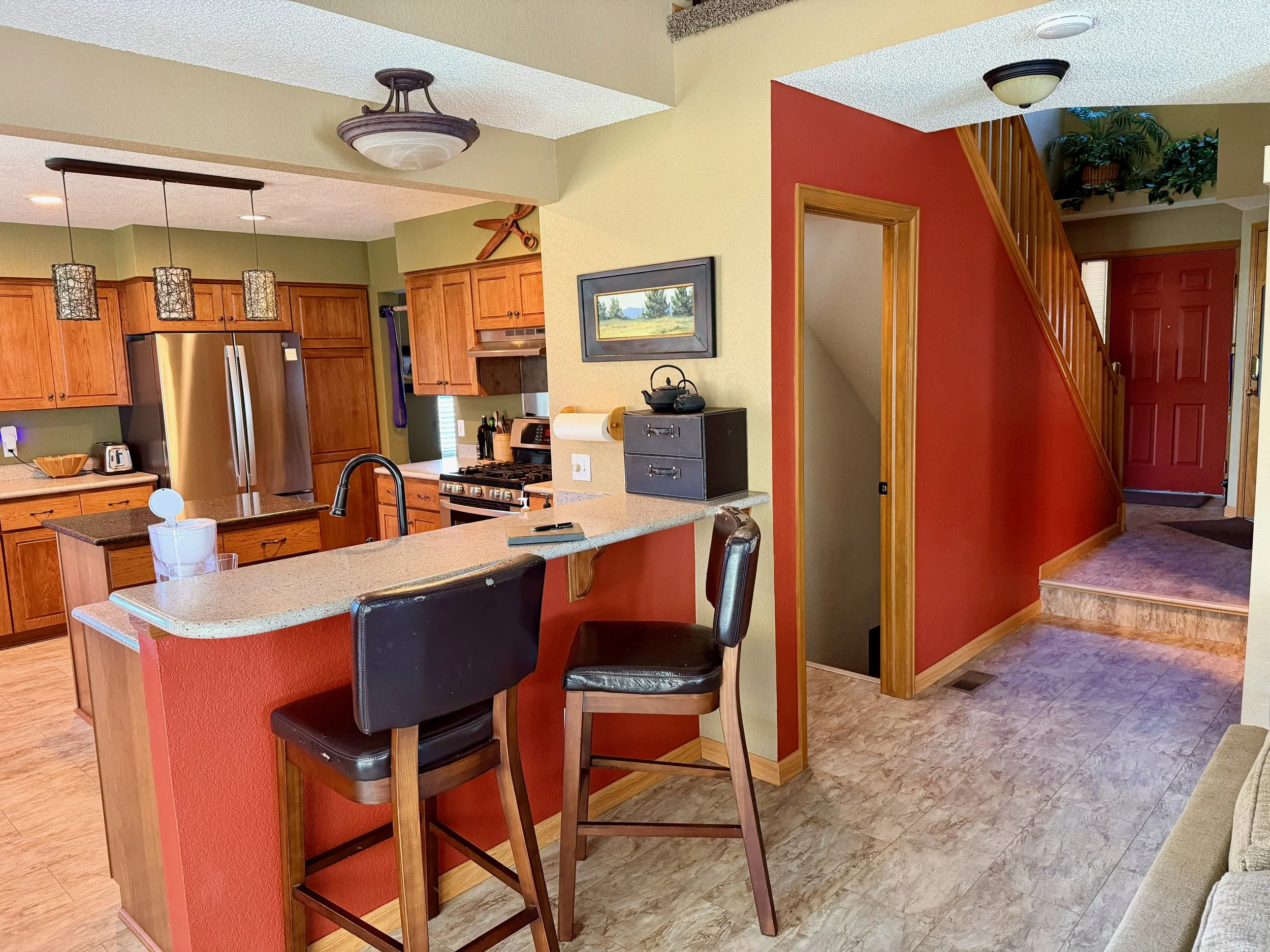 Before view of a client's kitchen and dining area with wooden cabinets, stainless steel refrigerator, black chairs, and a red accent wall.