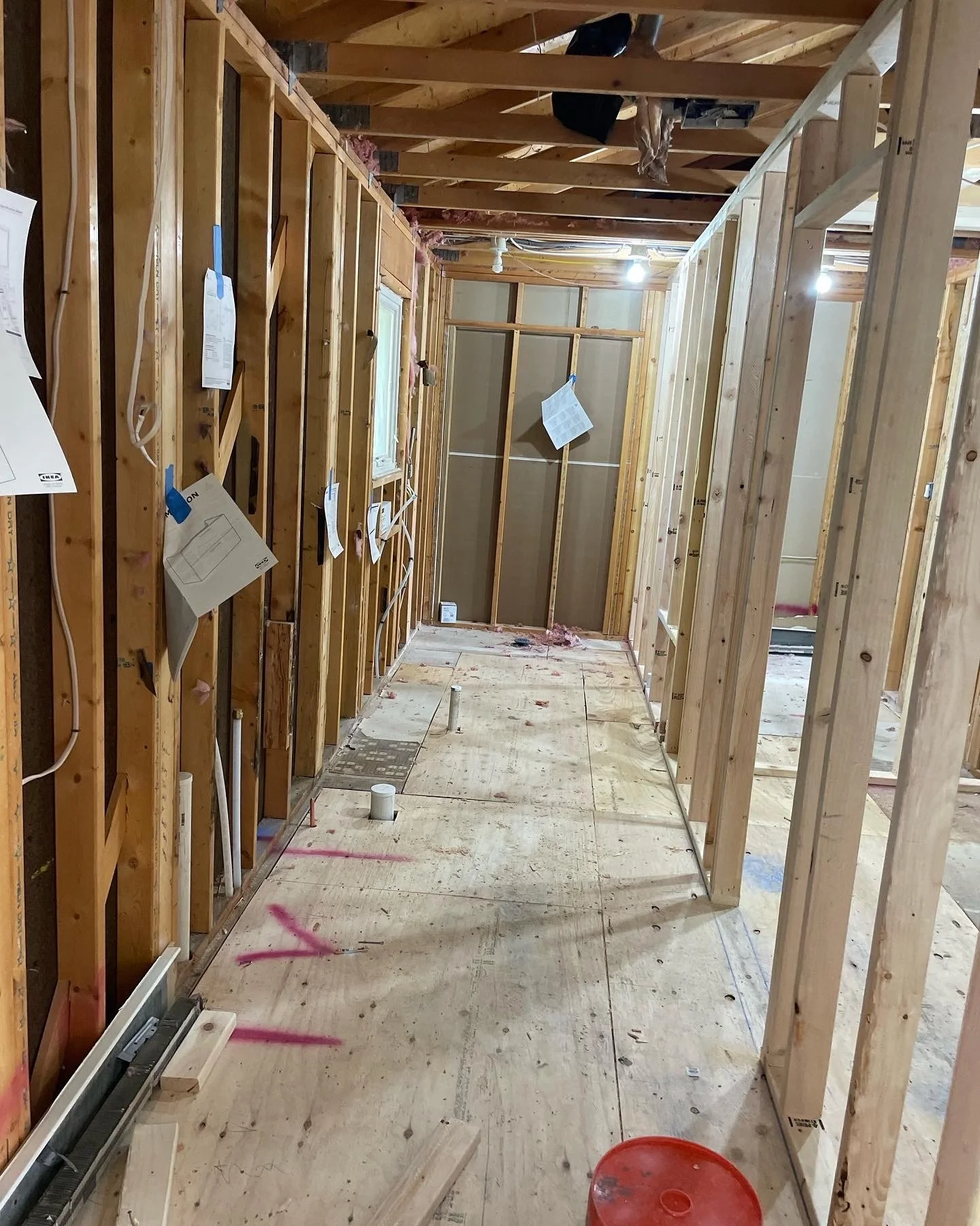 Interior view of a house under construction with exposed wooden framing, electrical wiring, and some construction materials on the plywood floor.