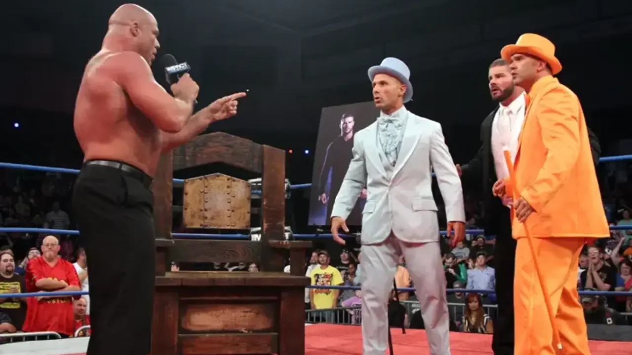 Kurt Angle pointing and speaking into a microphone across a chair in the ring toward Christopher Daniels, Frankie Kazarian, and Robert Roode during a TNA Wrestling segment.