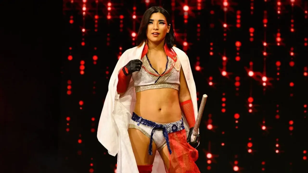 Professional wrestler Hikaru Shida standing on the AEW entrance stage wearing white and red gear with a white cape, holding a kendo stick against a background of flashing red lights.
