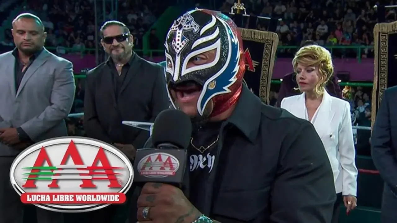 Rey Mysterio speaking into a Lucha Libre AAA microphone to announce upcoming matches for the Verano de Escándalo event.