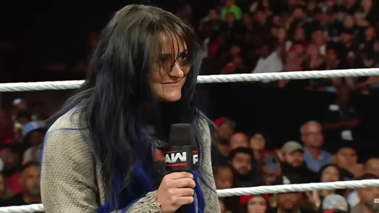 Lyra Valkyria on WWE Raw holding a microphone, sporting messy dark blue hair, a drab grey cardigan, and a wide, unsettling smile during her heel turn segment.