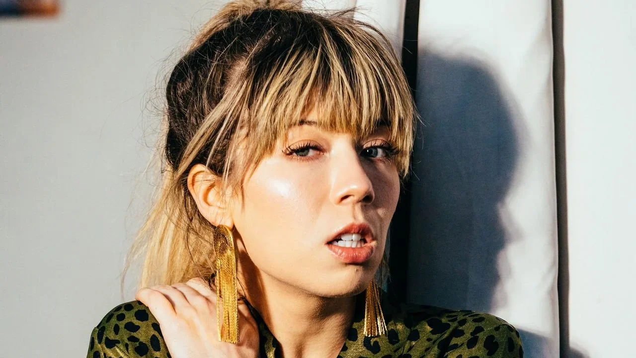 A portrait of Jennette McCurdy with blonde hair, bangs, and long gold fringe earrings, wearing a green animal-print top with her hand touching her opposite shoulder.