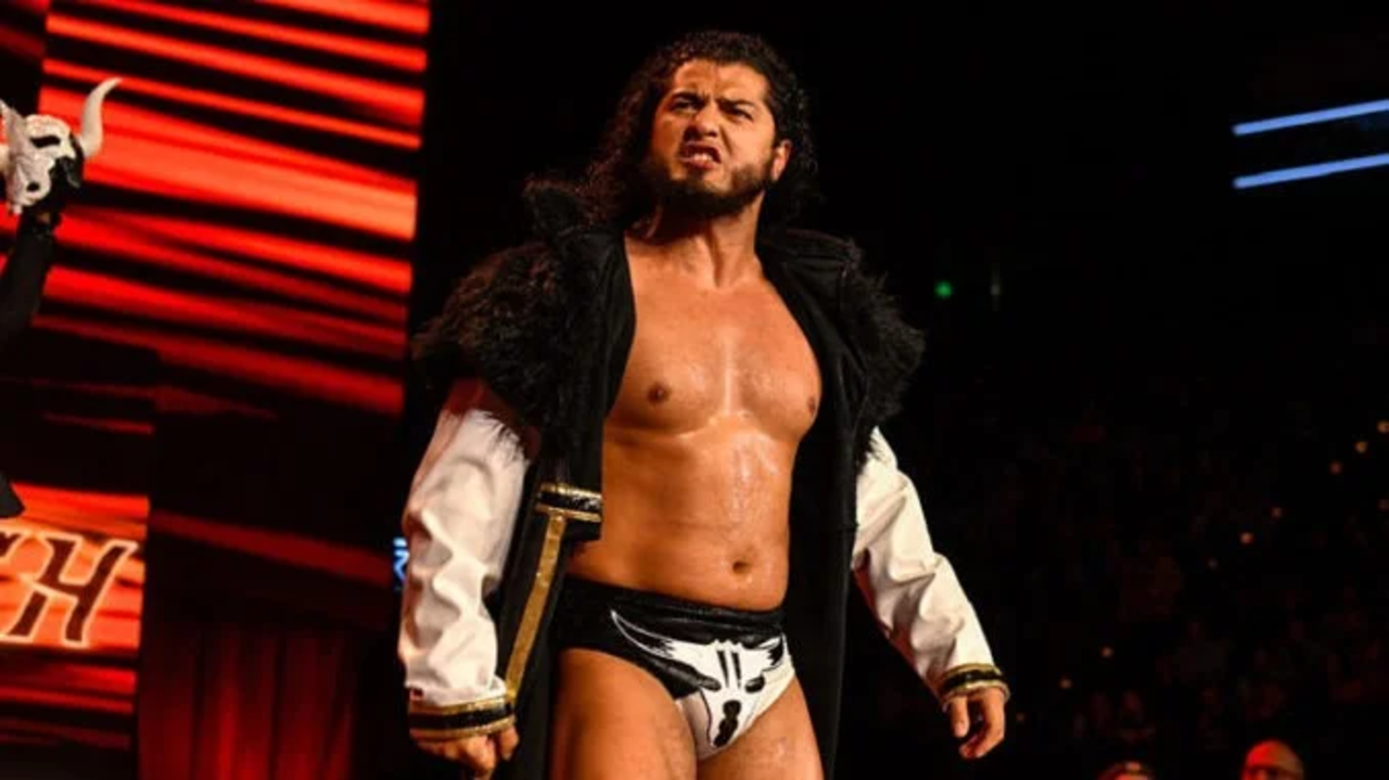 Professional wrestler RUSH standing in the ring during an AEW entrance, wearing his signature gear and jacket.