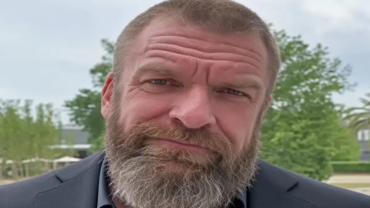 Triple H (Paul Levesque) with a short buzz cut and full salt-and-pepper beard, wearing a dark gray suit jacket over a dark button-up shirt, looking at the camera outdoors.