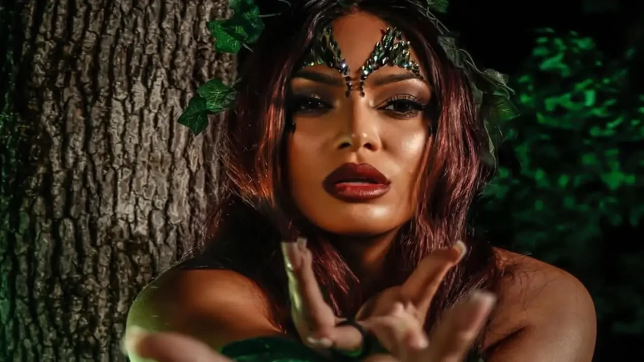Karissa Rivera posing outdoors in Poison Ivy-inspired makeup with dark green jewels on her forehead, ivy leaves in her hair, and a tree trunk in the background.