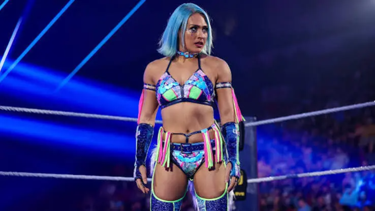 Giulia in a blue and pink patterned wrestling ring attire with bright blue hair, standing in the ring under arena lights.