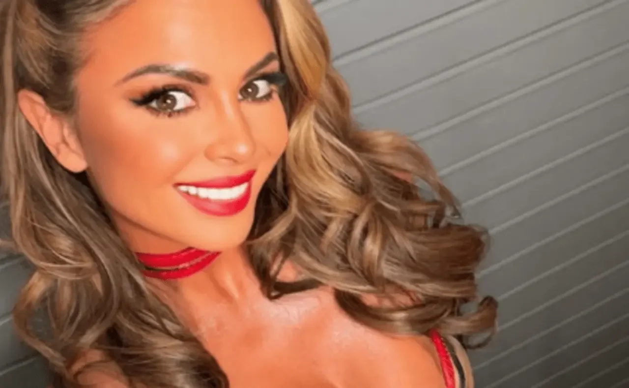 A close-up portrait of a smiling Harley Cameron with vibrant red lipstick and wavy blonde hair, looking toward the camera. She is wearing a red choker necklace and a matching red top against a neutral, grey textured background.