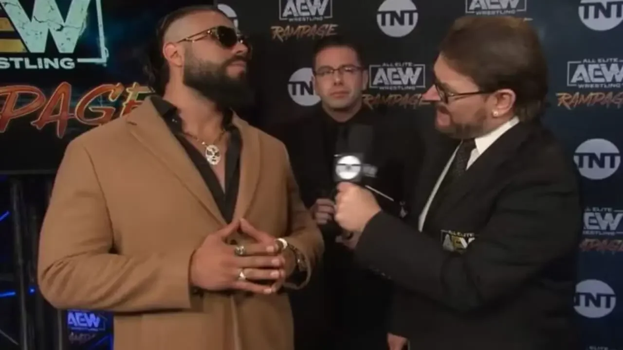 Andrade El Idolo wearing a tan jacket and sunglasses during an AEW backstage interview, looking cool and composed while being interviewed in front of an AEW Rampage backdrop.
