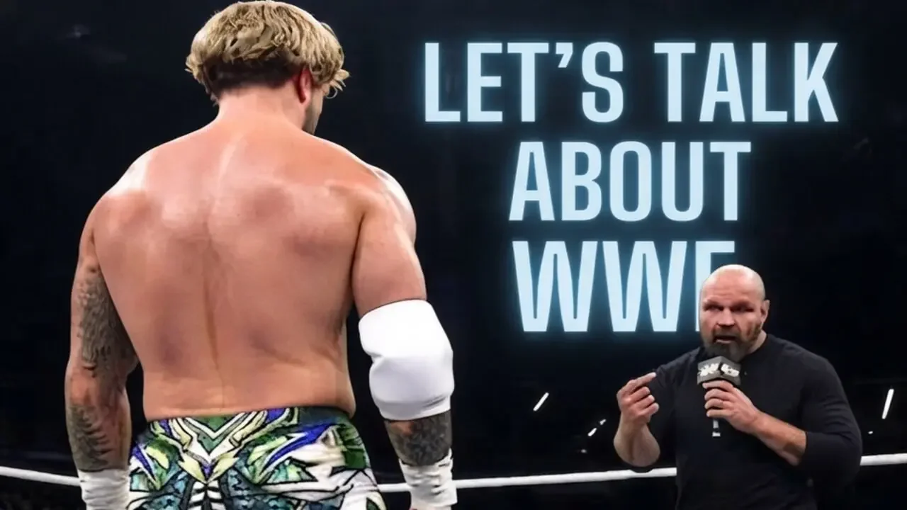 A photograph taken inside a professional wrestling ring, viewed from behind wrestler Will Ospreay (back to camera, detailed green and white gear) as he faces another wrestler (facing camera, bald, black shirt) holding a microphone.