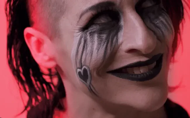 Close-up profile of professional wrestler Rosemary grinning maniacally under red lighting, wearing dark face paint, white contact lenses, dark lipstick, and a black heart emblem drawn on her cheek.