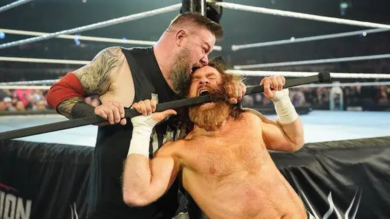 Kevin Owens choking Sami Zayn across his face and throat using a black Kendo stick against the ring post during a WWE match.