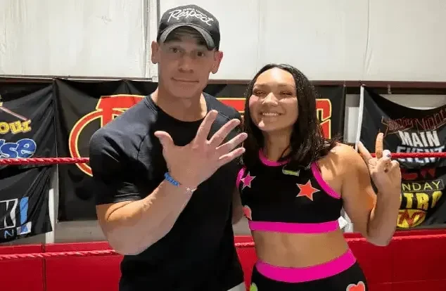 John Cena and Izzy smiling together inside a wrestling ring, with Cena holding up his hand to the camera and banners hanging in the background.