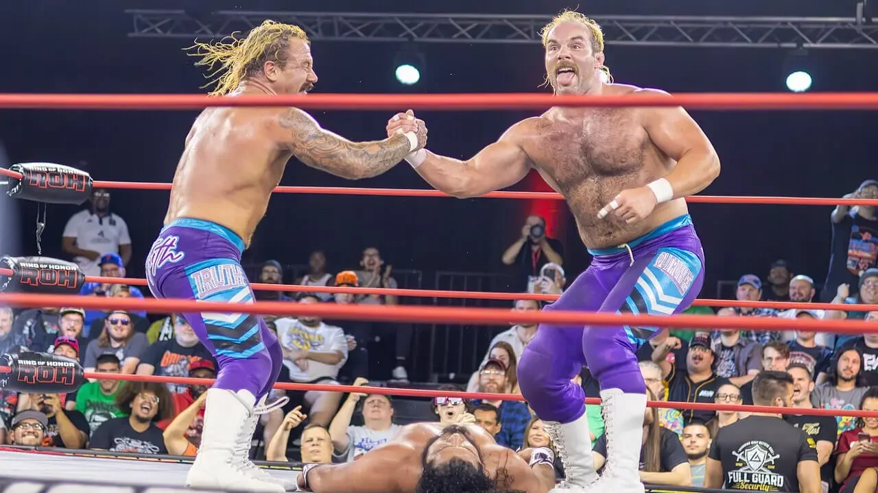 Professional wrestling tag team The Outrunners (Truth Magnum and Turbo Floyd) posing together in a wrestling ring, wearing matching purple and teal tights with white boots, while an opponent lies on the mat below them.