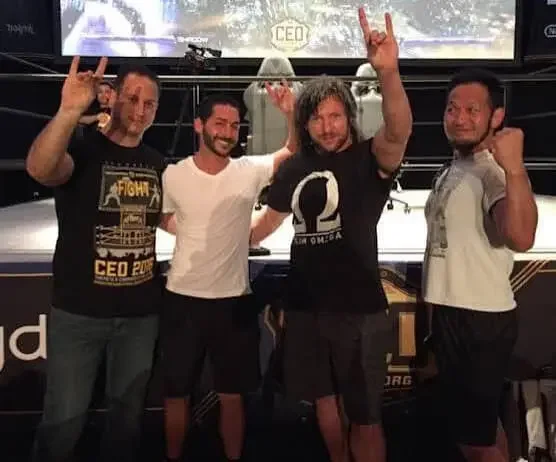 A 2018 photograph taken in a wrestling ring featuring four men posing together, including Kenny Omega and Chasyn Rance, with their hands raised in a gesture