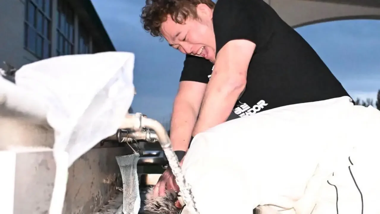 A professional wrestler in a black shirt and white pants aggressively scrubbing another person's head under a running water tap at an outdoor school wash station.