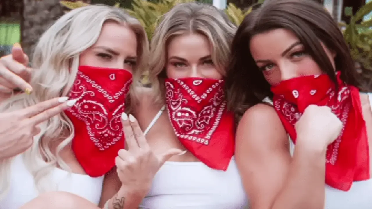 Close-up portrait of Ash by Elegance, Paige VanZant, and Mandy Rose wearing white tops and red bandanas covering the lower half of their faces while posing together outdoors.