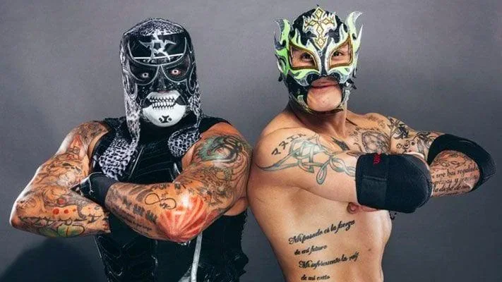 The Lucha Bros (Penta and Fenix) posing back-to-back with arms crossed wearing their signature wrestling masks and gear.