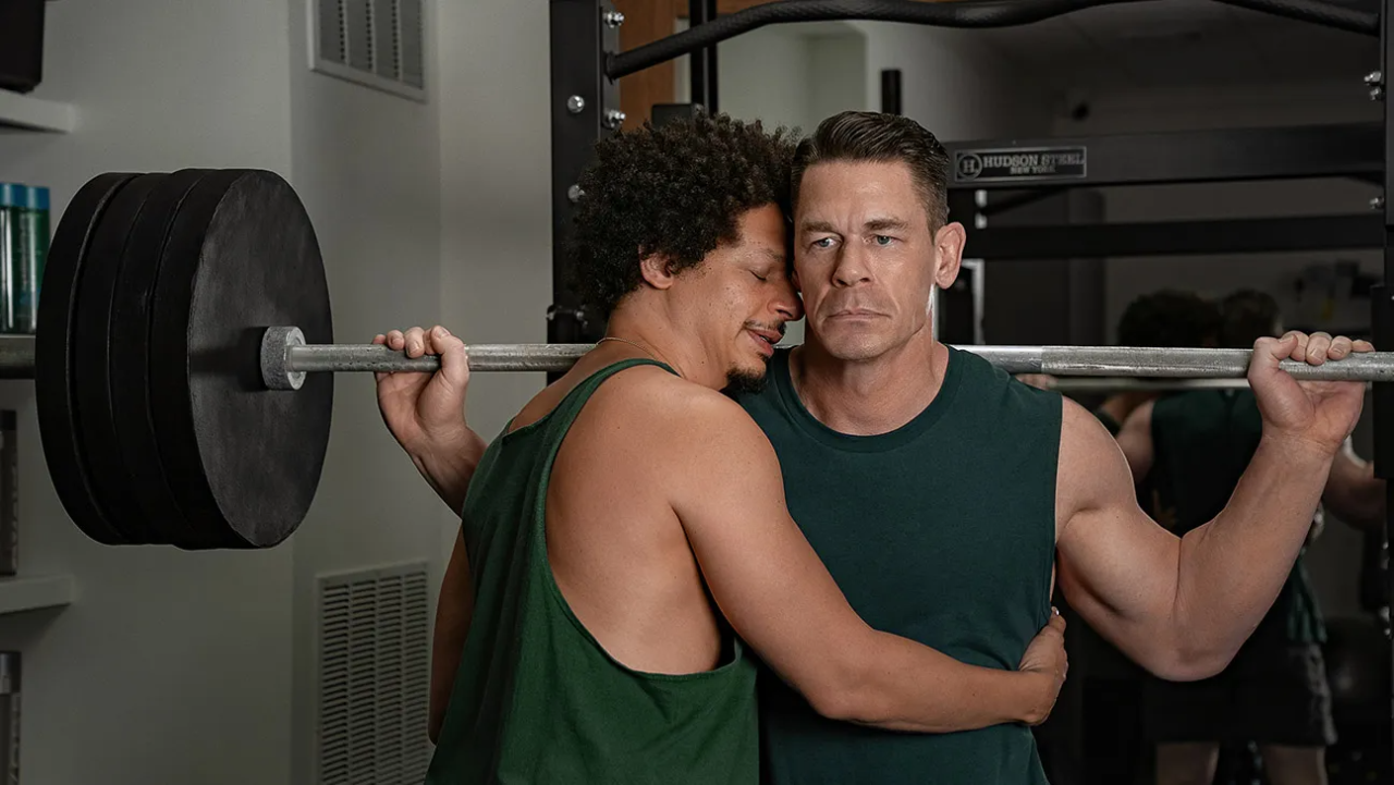 Eric André with his eyes closed hugging a stoic John Cena as Cena holds a heavily loaded barbell on his shoulders in a gym squat rack from the Netflix movie Little Brother.