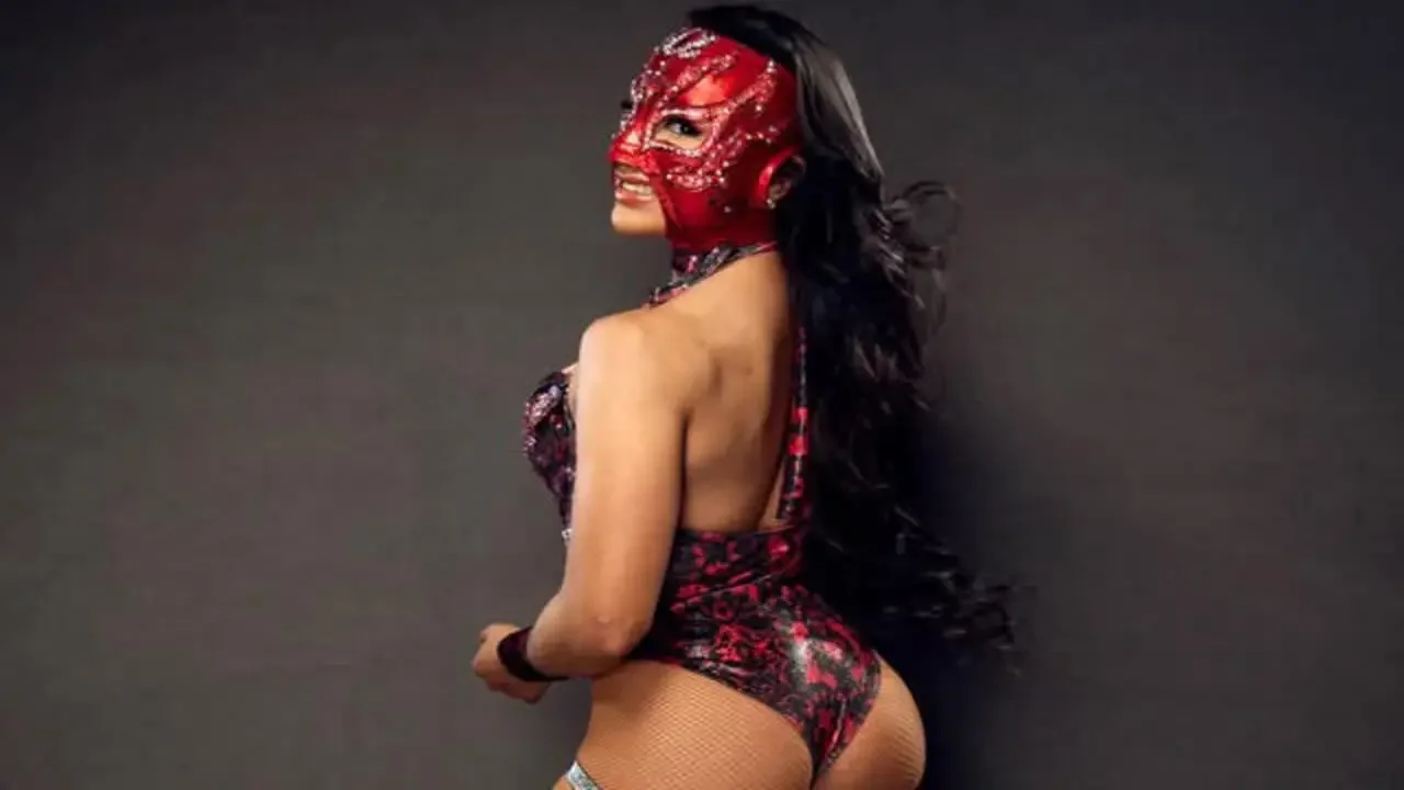 Deli Cious Lily wearing a red and black patterned wrestling outfit and a matching mask while posing against a dark studio background.