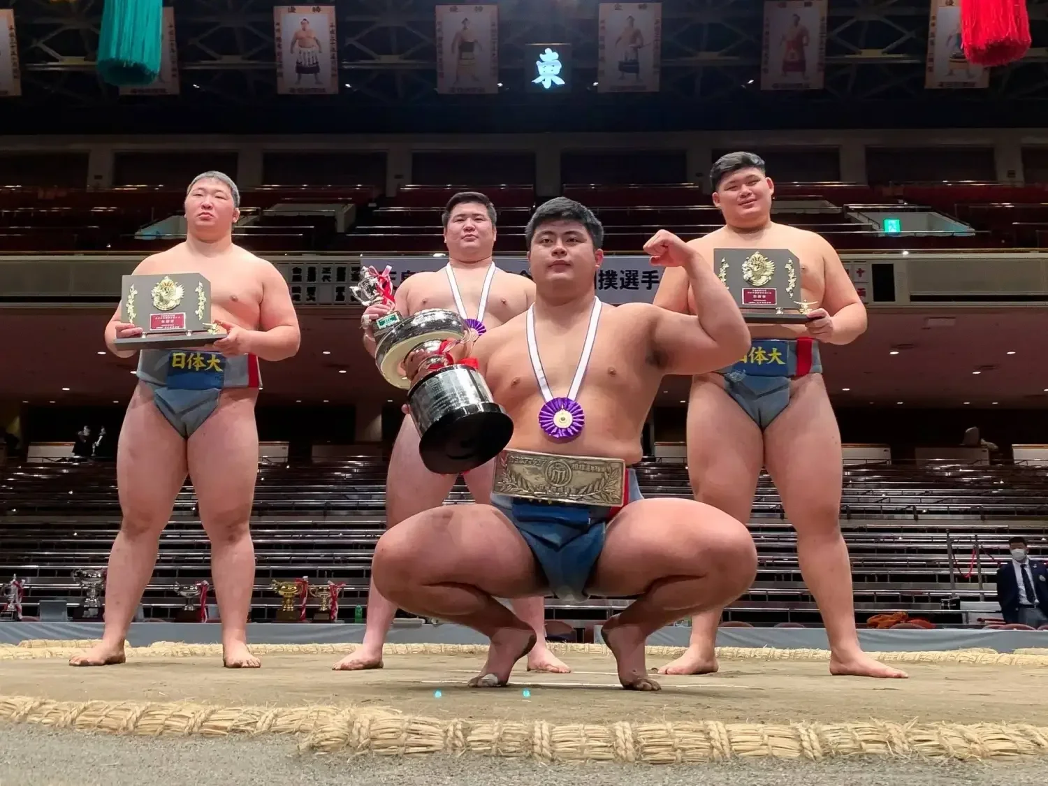 Hidetora Hanada posing on a sumo ring holding a trophy and wearing a medal.
