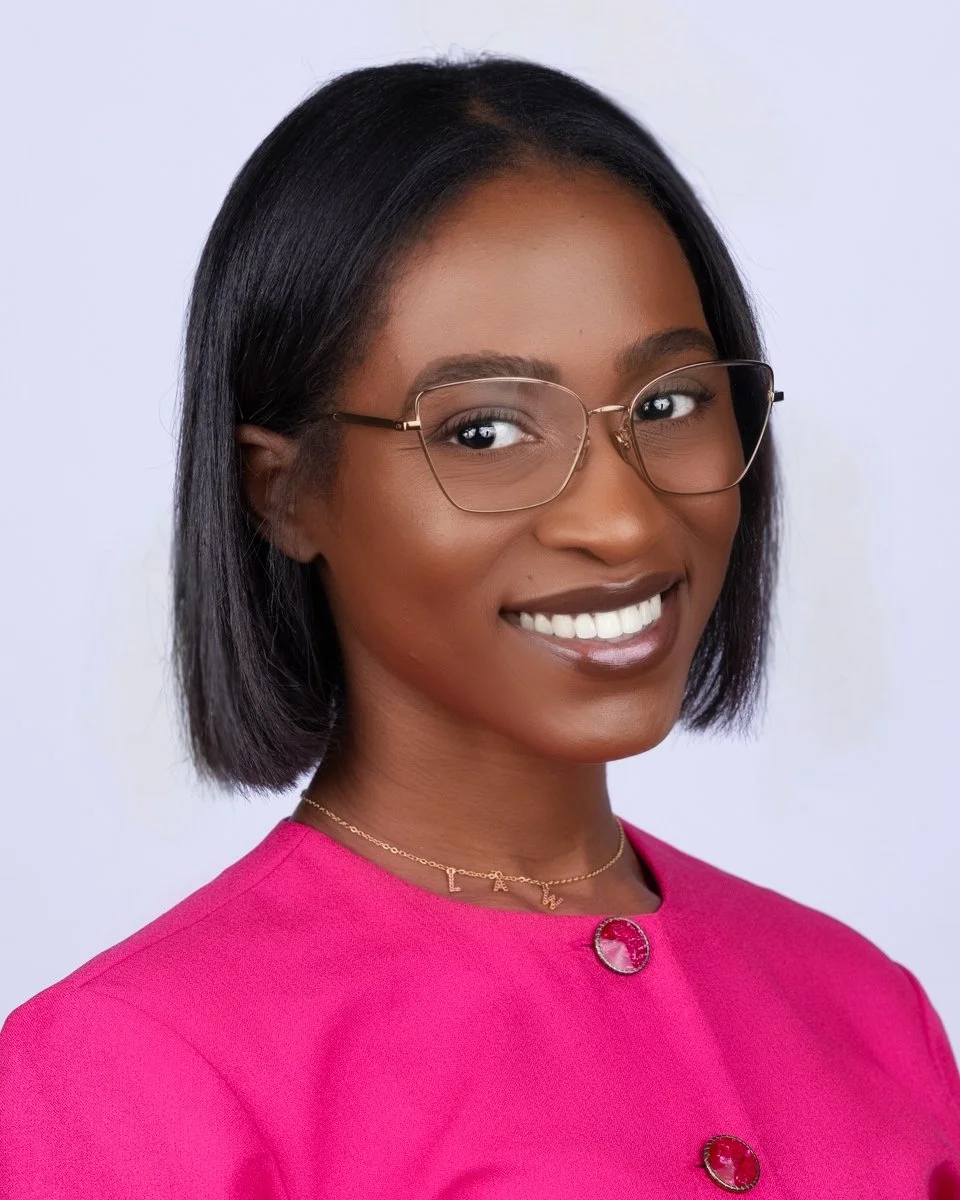 A young woman with short black hair, wearing glasses, a pink top with decorative buttons, and a delicate necklace, smiling at the camera against a plain white background.
