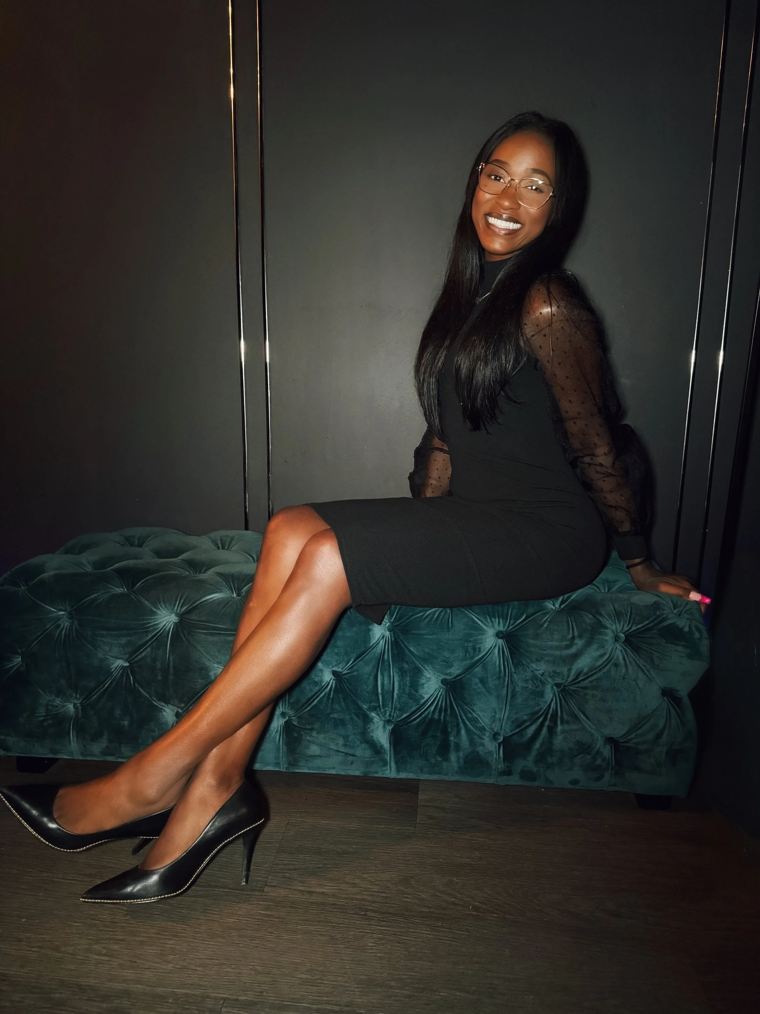 A woman with long black hair and glasses, smiling, wearing a black dress with sheer polka-dot sleeves, sitting on a teal velvet tufted bench in a dark room.