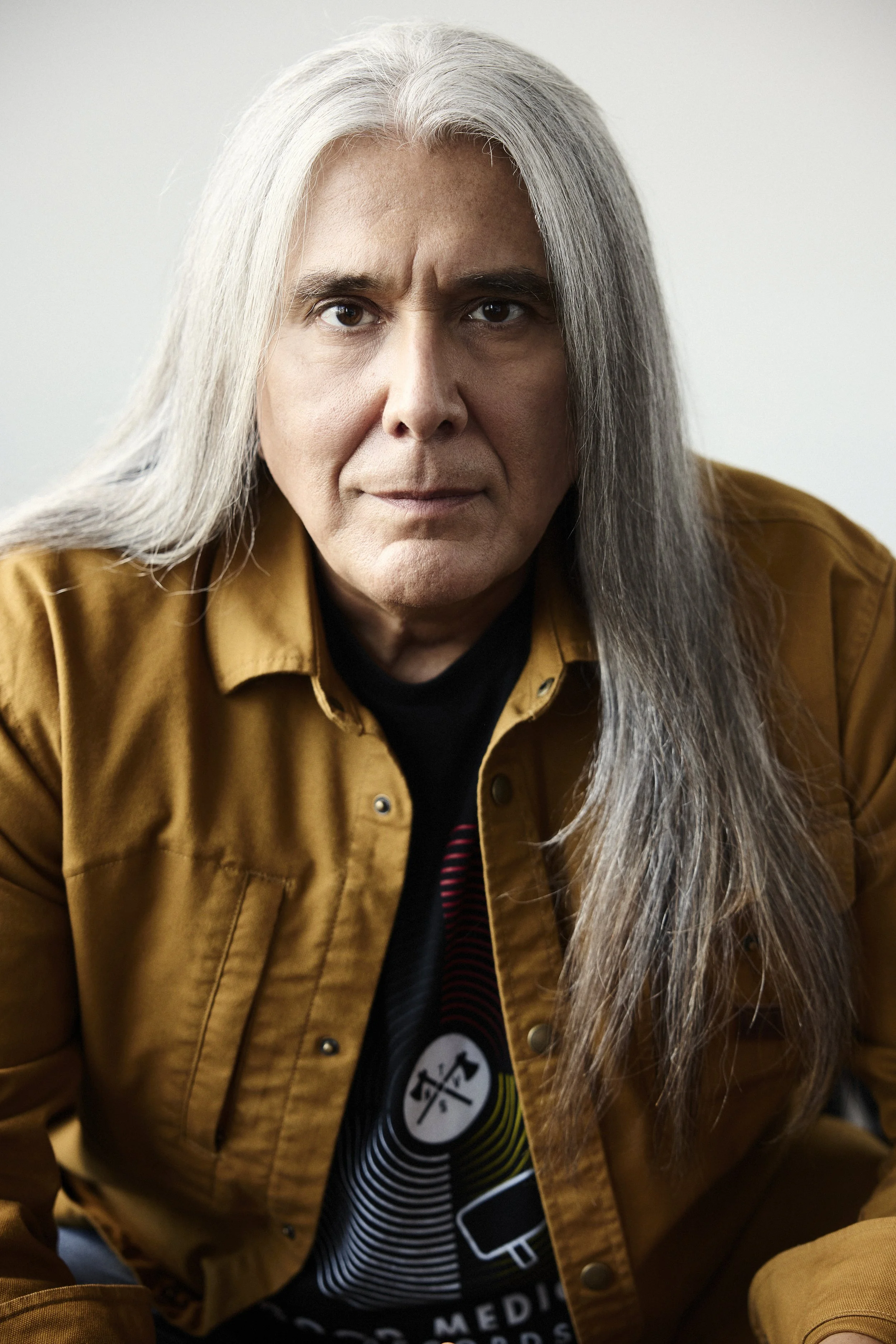 Close-up of a man with long gray hair, wearing a brown jacket and a black t-shirt with a graphic design, sitting indoors against a plain background.