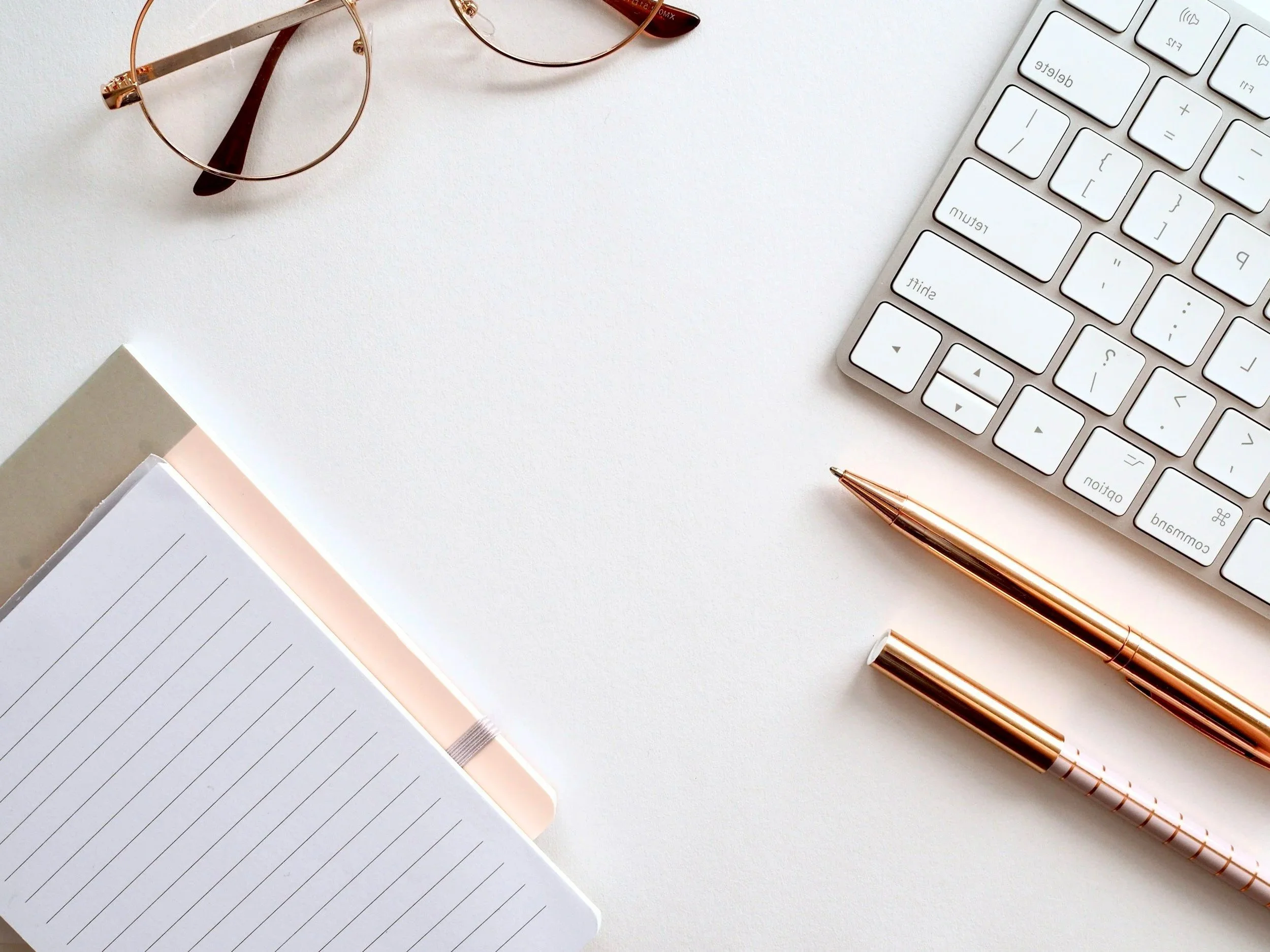 Top view of a white desk with a pair of eyeglasses, a white keyboard, two rose gold pens, and a lined notebook.