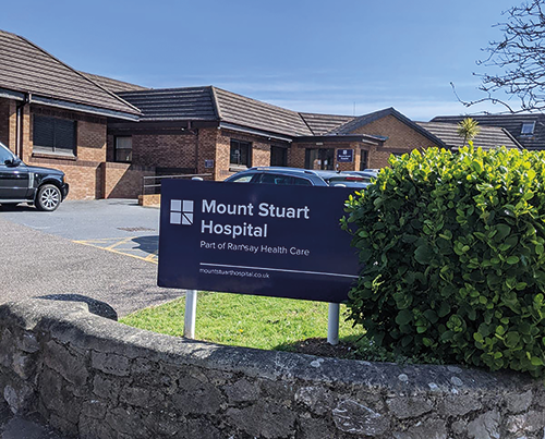 Sign for Mount Stuart Hospital, part of Ramsay Health Care, outside a brick building with parked cars and a bush in the foreground.