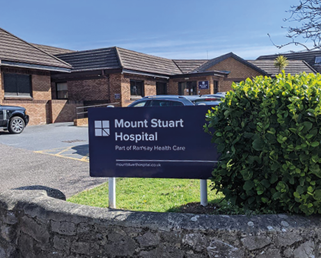 Sign outside Mount Stuart Hospital with parking lot and brick buildings in the background.