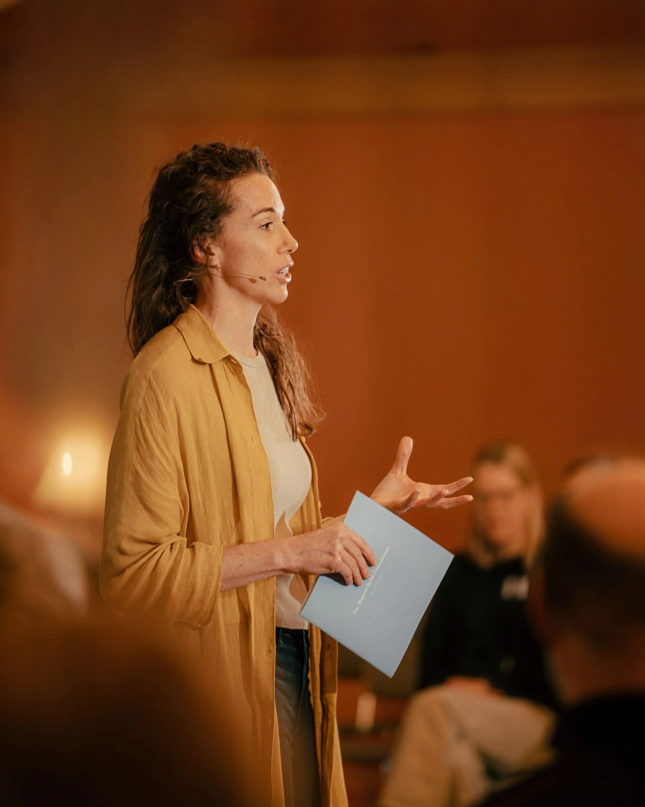 Kyra Spira giving a speech or presentation, holding a folder or paper, in a room with warm lighting and blurred audience members in the background.