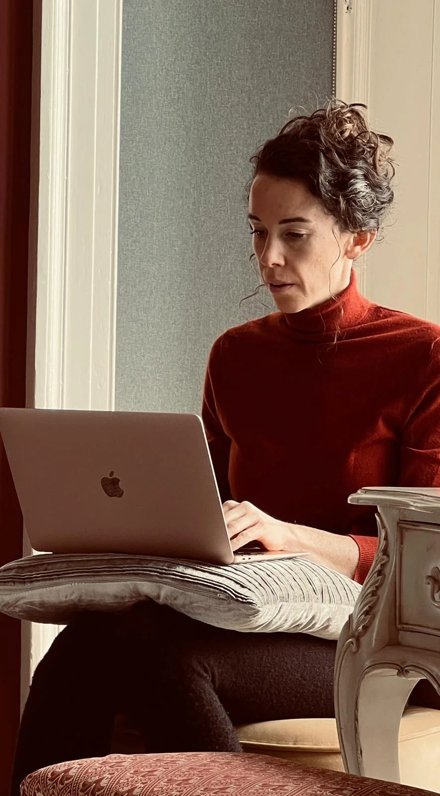 Kyra Spira sitting at a vintage-style desk, working on a MacBook, with a gray wall behind her.