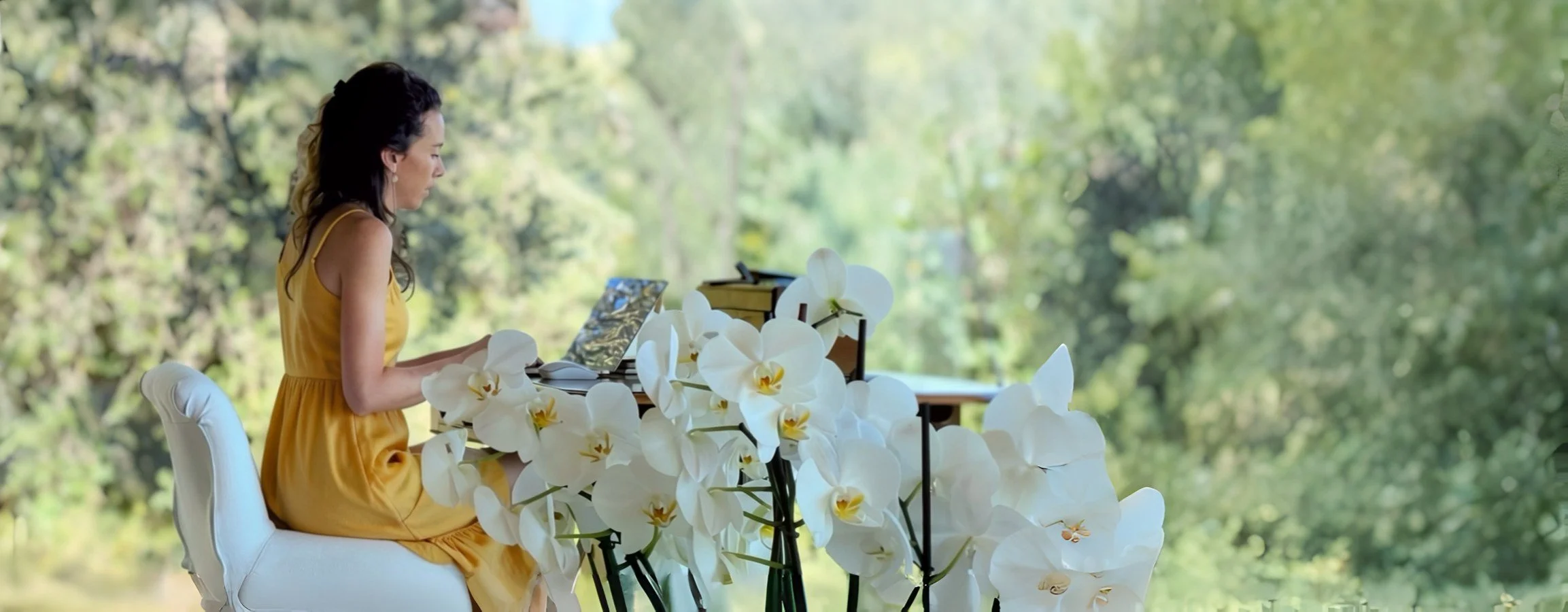 Kyra Spira sitting at a desk decorated with white orchids with a lush green forest background.