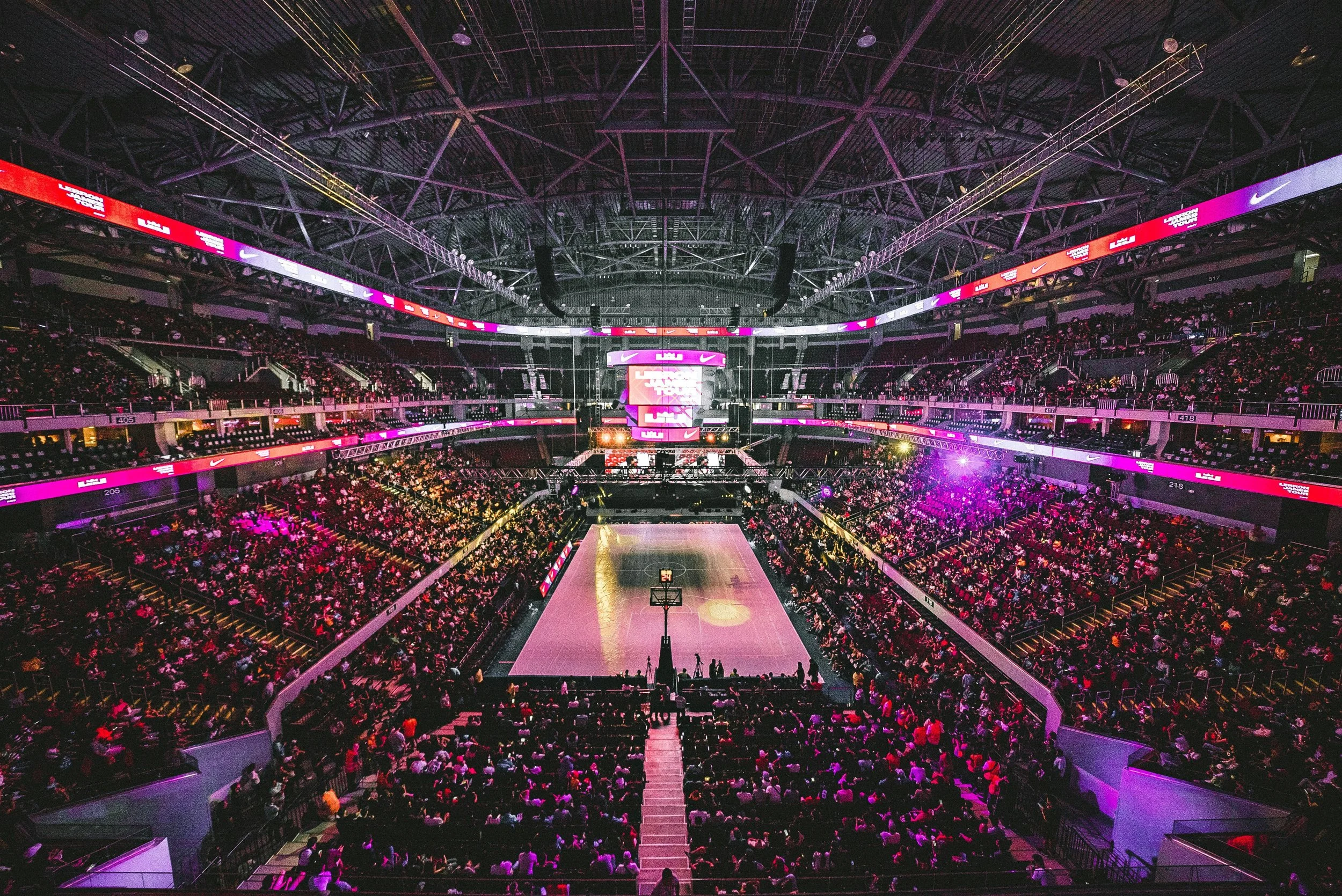 Indoor basketball arena filled with spectators, illuminated in pink and purple lights, with the basketball court in the center and a large digital scoreboard hanging from the ceiling.
