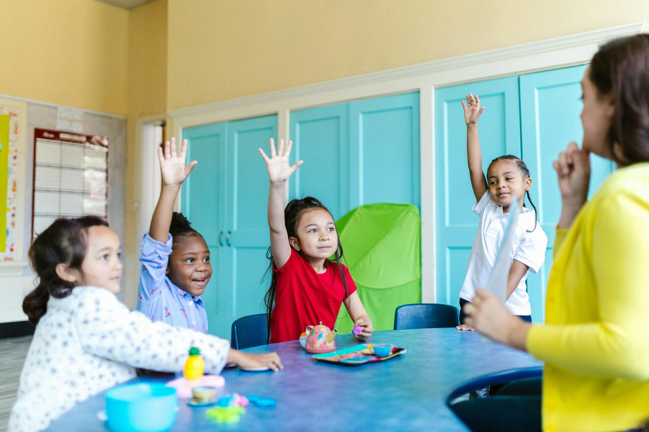 Children at a classroom table with some raising hands to answer, an adult teacher on the right observing.