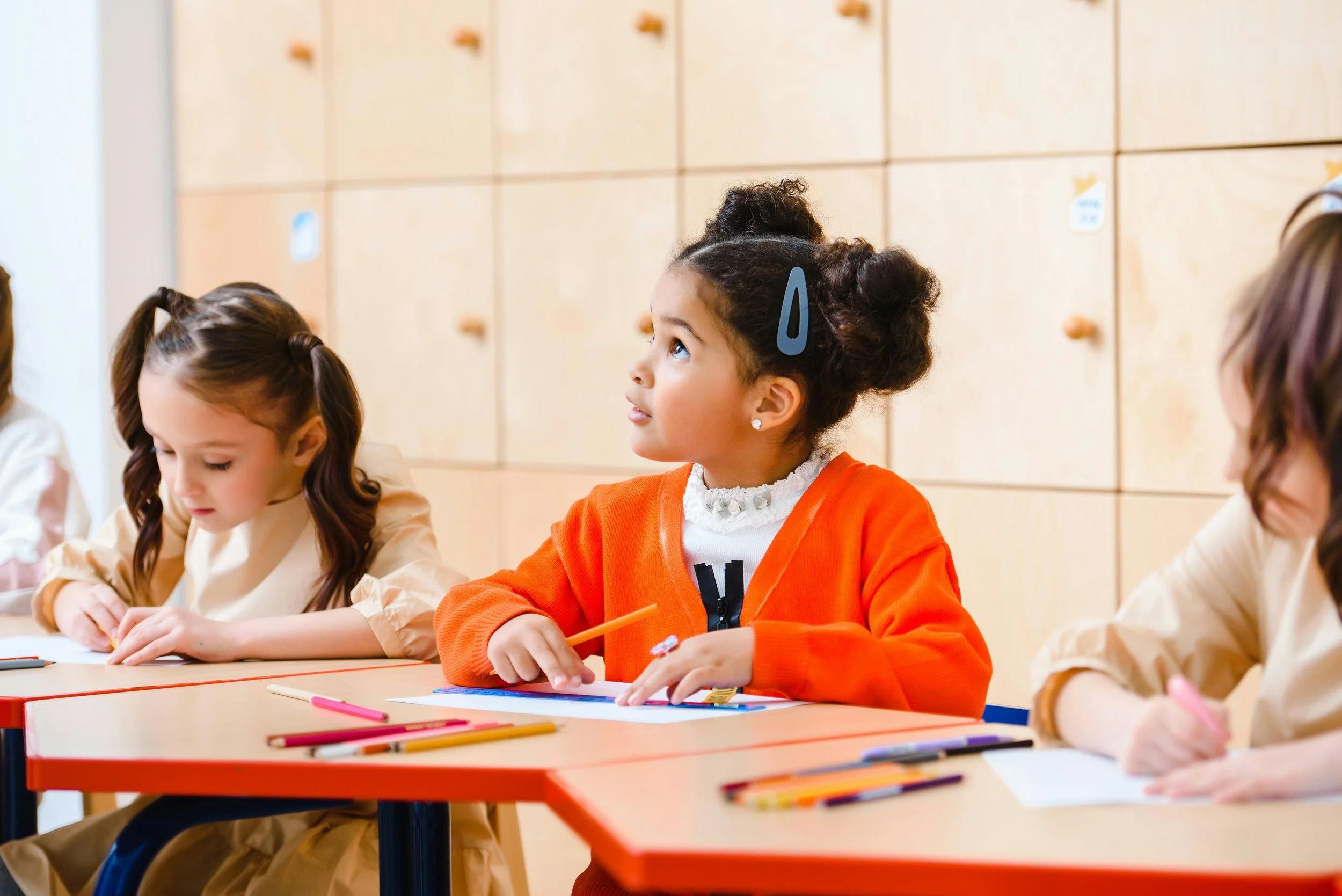 A group of schoolchildren sitting at desks in a classroom, with one girl in an orange sweater appearing attentive and looking up while others are focused on their work.