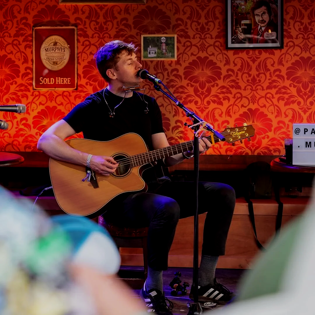 A young man with a guitar, singing into a microphone on a stage with red floral wallpaper and framed pictures behind him.