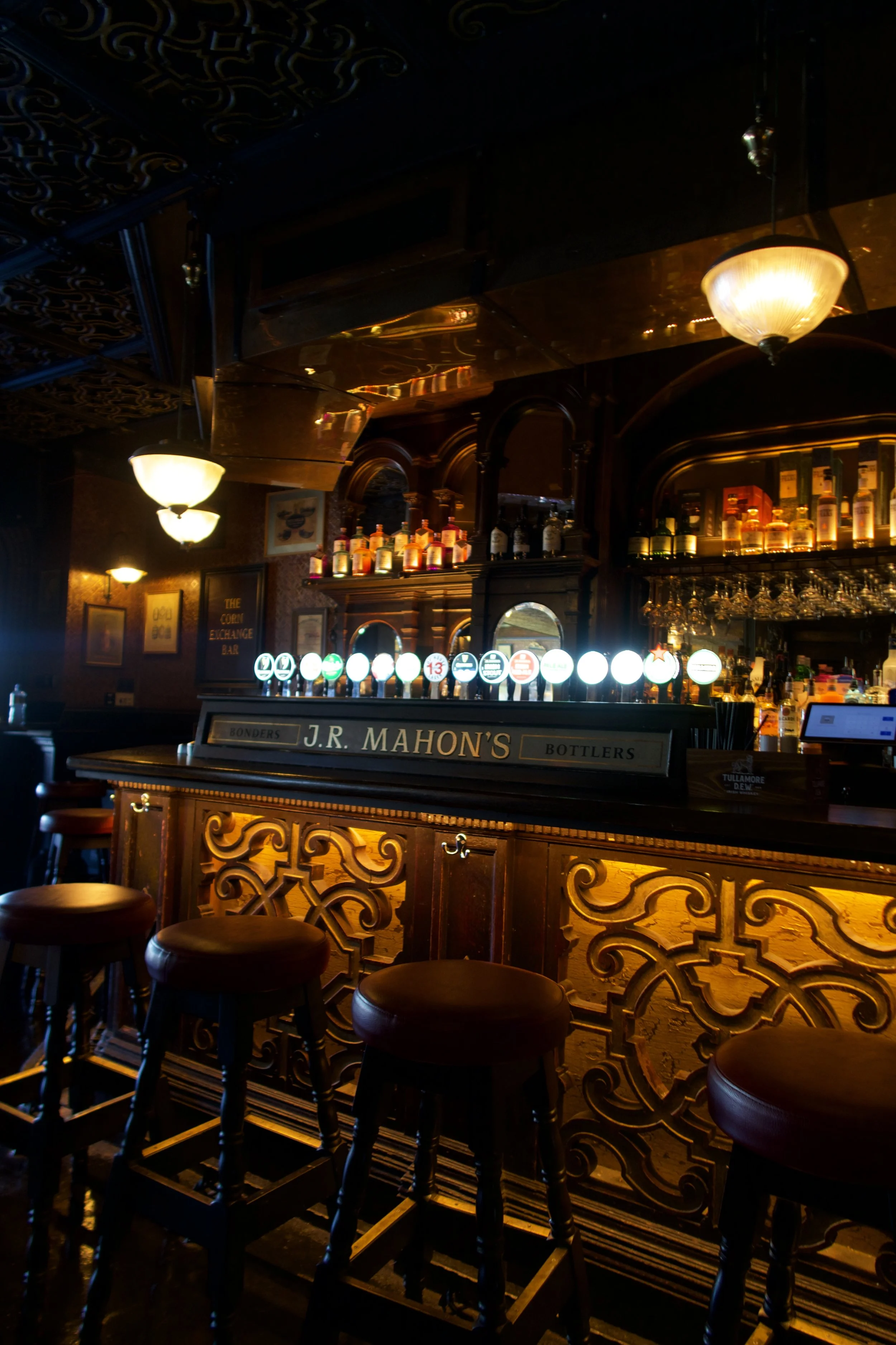 Interior of a dimly lit bar with three golden pendant lights hanging from the ceiling and two large windows showing a brick building outside.