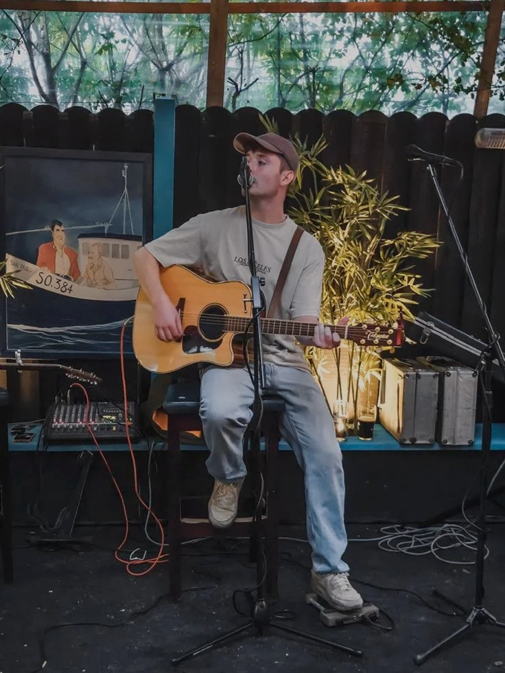 Young man playing an acoustic guitar and singing at a small outdoor music setup with plants and a black wooden fence in the background.