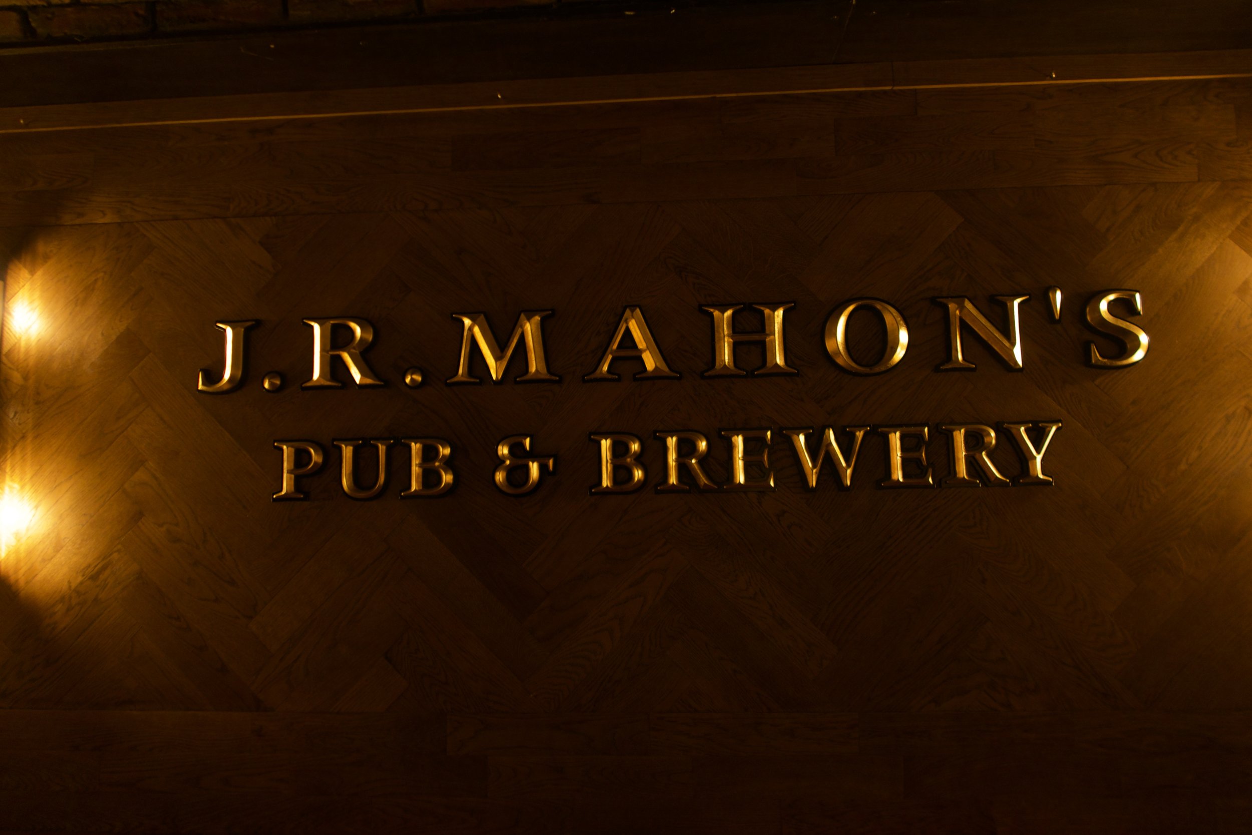 Interior of a dimly lit restaurant or bar with black walls, gold accents, and modern lighting fixtures. There are yellow booths along the wall and chairs around tables, with some windows visible in the background.