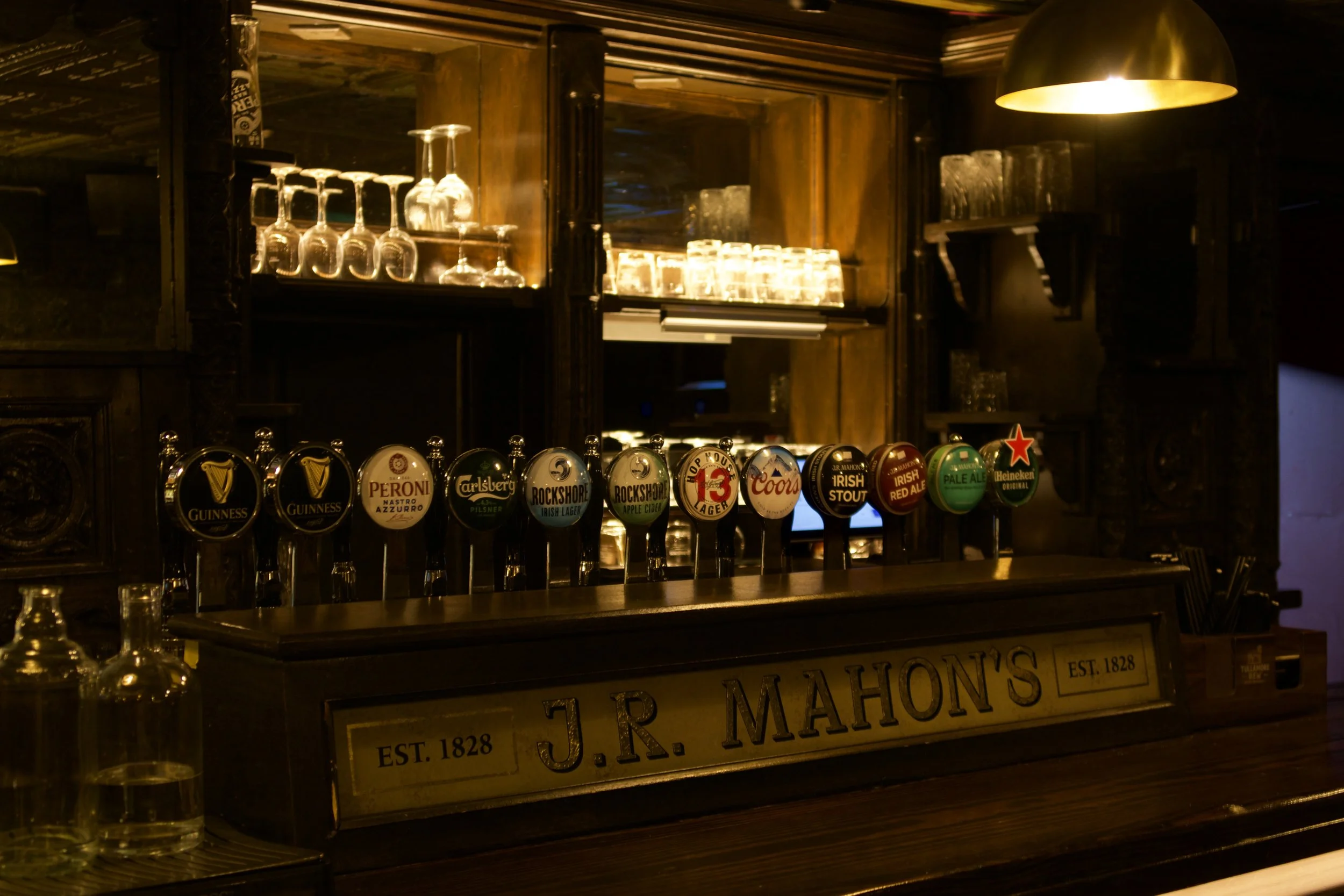 A dimly lit bar with a wooden counter, bar stools, and shelves stocked with various bottles of alcohol. There are TV screens displaying digital content behind the bar.